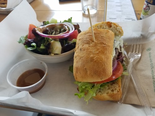 A sandwich with lettuce, tomato, and onion, served with a side salad and a small cup of dressing on a tray.