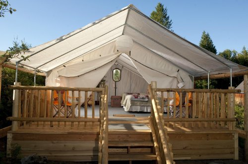 A spacious glamping tent with wooden steps, featuring a cozy interior and outdoor seating. Surrounded by trees.