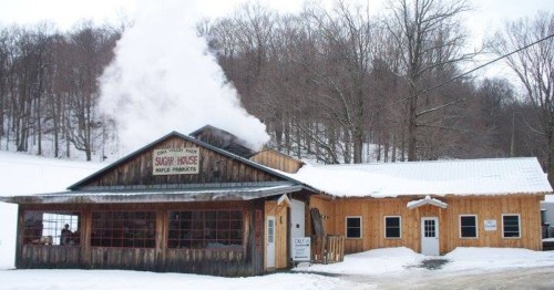 A rustic sugar house in winter, with smoke rising from the chimney and snow covering the ground.