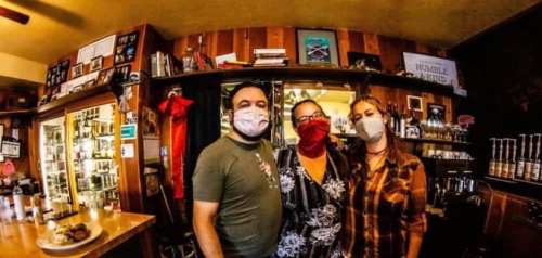 Three people wearing masks stand together in a cozy bar, surrounded by shelves and decor.