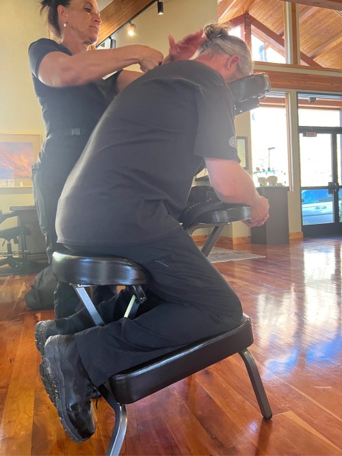 A person receives a seated massage in a bright, wooden-floored room with large windows.
