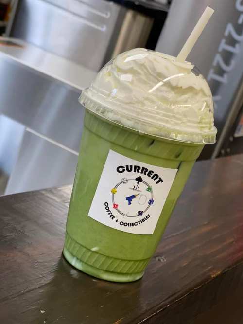 A green iced drink with whipped cream in a clear cup, featuring a logo for "Current Coffee + Collectibles."
