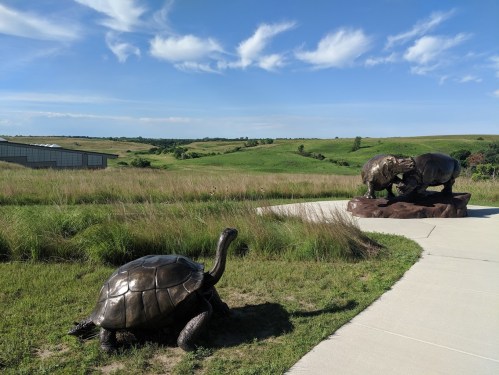 A bronze turtle and a bear sculpture on a pathway, surrounded by green hills and a blue sky with clouds.