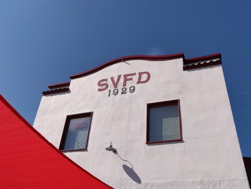 White building with "SVFD 1929" on the facade, blue sky above, and a red awning in the foreground.