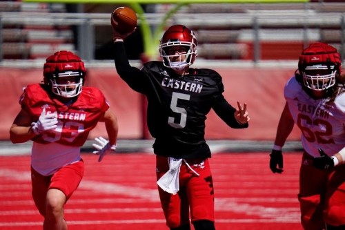 A football player in a black jersey throws a pass while two teammates in red jerseys run nearby on a field.