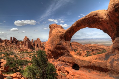 A stunning view of a natural rock arch surrounded by red rock formations and a blue sky with scattered clouds.
