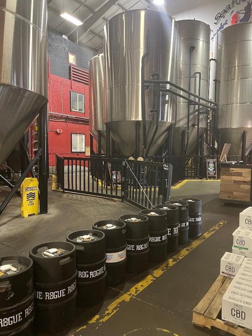 Interior of a brewery featuring large stainless steel tanks and barrels lined up along the floor.
