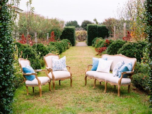 A serene garden path lined with hedges, featuring elegant chairs and a sofa adorned with blue and white pillows.