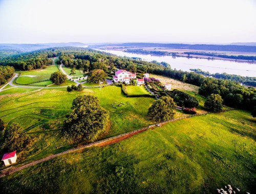 Aerial view of a lush green landscape with a river, featuring a large house and surrounding gardens.