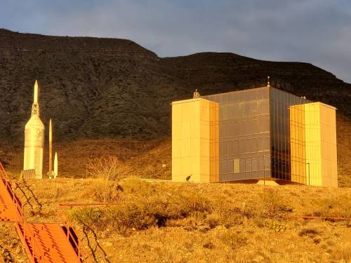 A modern building with large glass windows beside two rocket structures, set against a mountainous landscape at sunset.