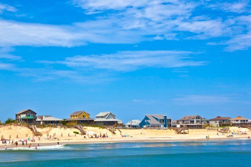 Colorful beach houses line a sandy shore under a bright blue sky, with people enjoying the beach and water.
