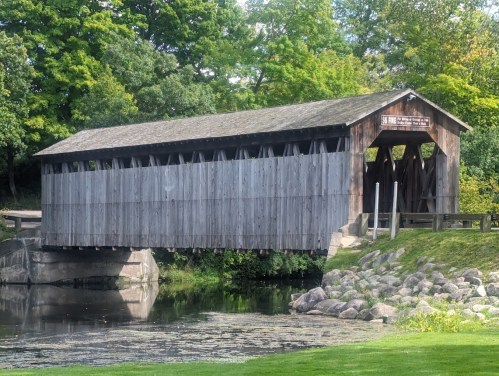 A wooden covered bridge spans a calm river, surrounded by lush greenery and rocks along the bank.