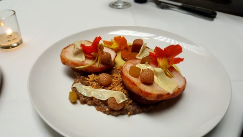 A gourmet dish featuring two halved fruits, garnished with flowers, nuts, and creamy elements on a textured base.