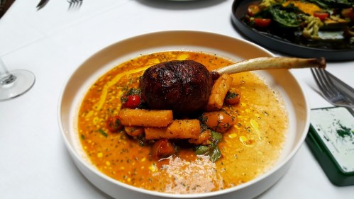 A gourmet dish featuring a roasted lamb chop on a bed of vegetables and sauce, garnished with herbs.