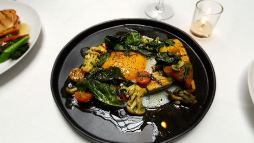A black plate with a sunny-side-up egg, sautéed vegetables, and herbs, garnished with cherry tomatoes and greens.
