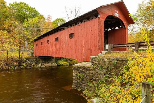 A red covered bridge spans a calm river, surrounded by autumn foliage and stone supports.