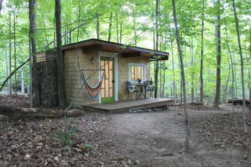 A small wooden cabin surrounded by trees, featuring a porch with a table and chairs, and a hammock nearby.