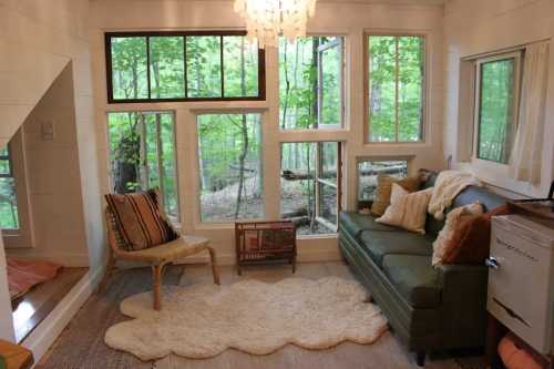 Cozy living space with large windows, green sofa, and a soft rug, surrounded by trees and natural light.