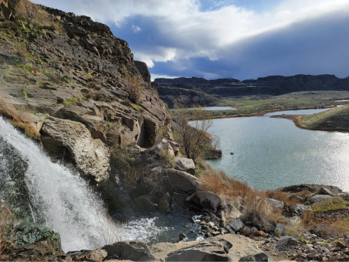A scenic view of a waterfall cascading into a calm lake, surrounded by rocky cliffs and green hills under a cloudy sky.