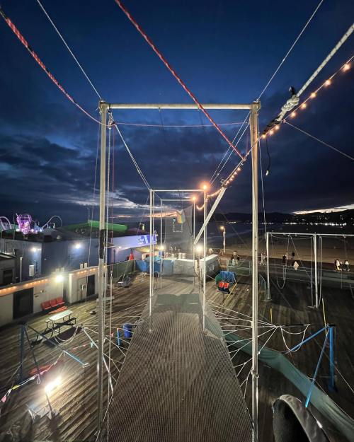 A view of a trapeze setup at dusk, with lights illuminating the area and a cloudy sky in the background.