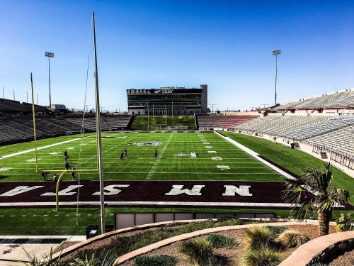 A wide view of a football stadium with green turf, goalposts, and empty stands under a clear blue sky.