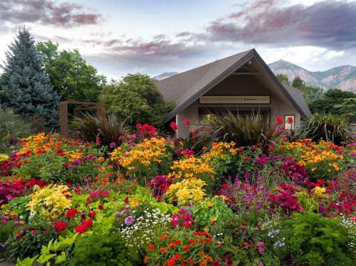 A vibrant garden filled with colorful flowers surrounds a modern building against a backdrop of mountains and a cloudy sky.