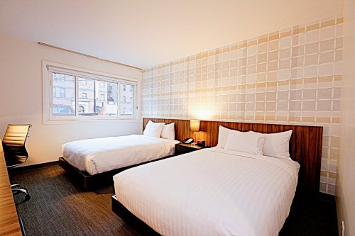 A modern hotel room featuring two beds, a desk, and a large window with light-colored walls and simple decor.