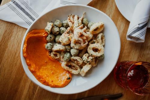 A bowl of fried calamari and green olives served with a side of orange dipping sauce on a wooden table.