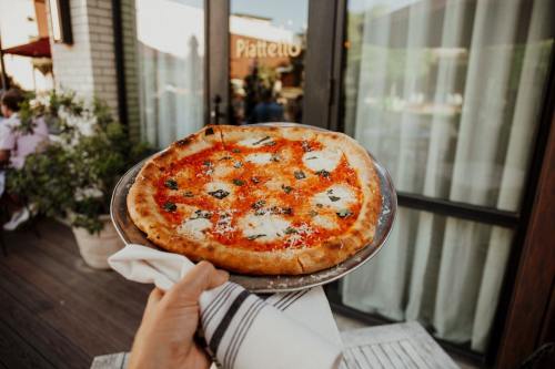 A hand holds a freshly baked pizza with cheese and basil on a silver platter outside a restaurant.