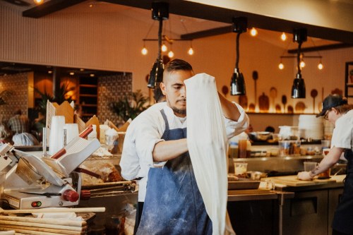 A chef in an apron prepares dough in a bustling restaurant kitchen with warm lighting and wooden decor.