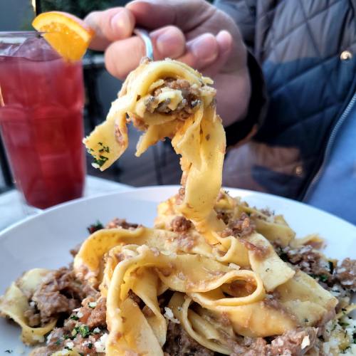 A hand holds a fork with fettuccine pasta topped with meat sauce, alongside a drink with an orange slice.