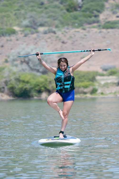 A person balances on a paddleboard in calm water, holding a paddle above their head with one leg raised.