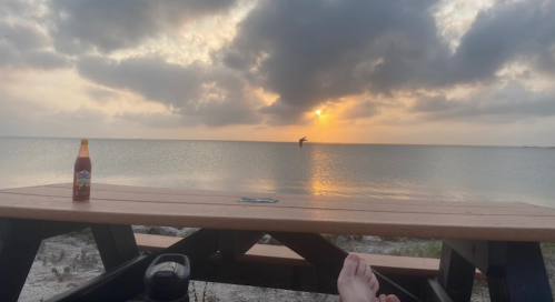 A serene beach scene at sunset, featuring a picnic table, a drink, and a person in the distance against the water.