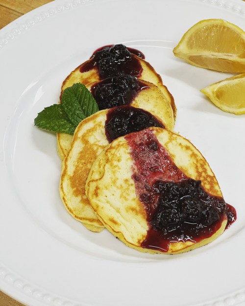 Three pancakes topped with berry jam, garnished with mint leaves and lemon wedges on a white plate.