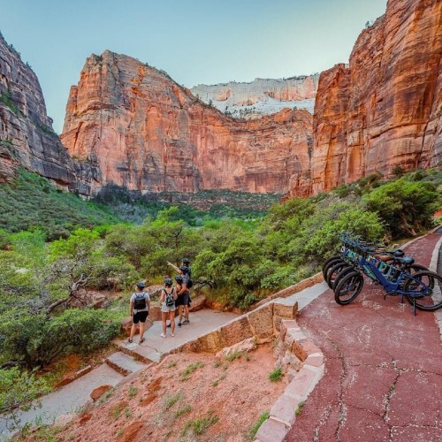 Hikers on a trail in a canyon surrounded by towering red rock formations and lush greenery. Bicycles parked nearby.