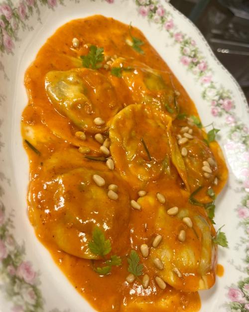 A plate of ravioli in a rich tomato sauce, garnished with pine nuts and fresh herbs.