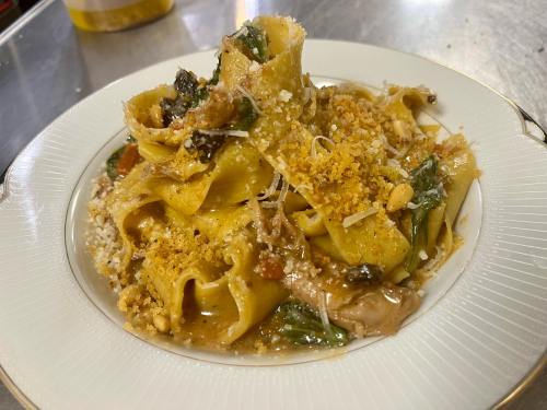 A plate of pappardelle pasta topped with mushrooms, greens, breadcrumbs, and grated cheese, served elegantly.