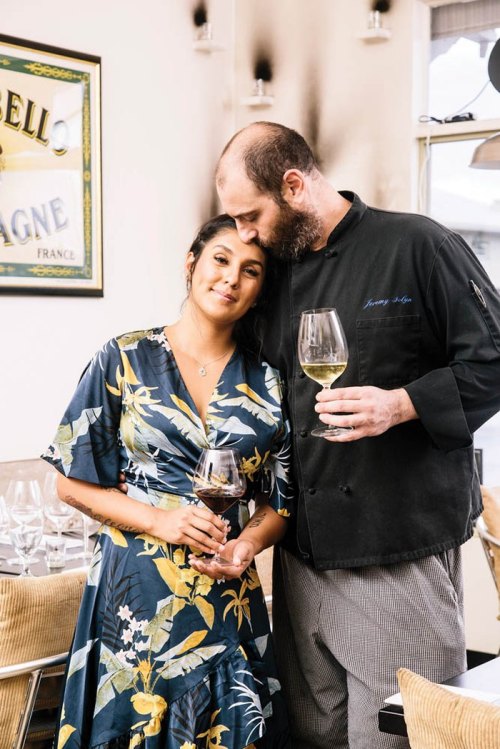 A couple stands together in a restaurant, holding wine glasses, with the man in a chef's coat and the woman in a floral dress.