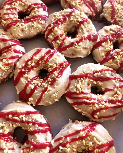 A close-up of donuts topped with creamy frosting, crushed peanuts, and red drizzle, arranged on a tray.