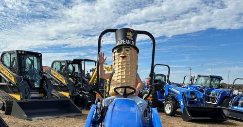 A person in a peanut costume waves from a blue tractor at a machinery event, surrounded by construction equipment.