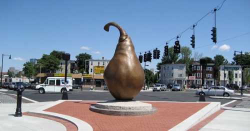 A large bronze pear-shaped sculpture stands in a plaza, surrounded by traffic lights and buildings under a clear blue sky.