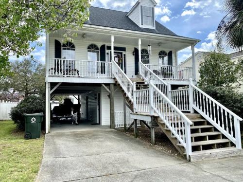A two-story house with a front porch, stairs, and a green trash bin in the driveway under a partly cloudy sky.