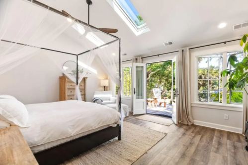 Bright bedroom with a four-poster bed, large windows, and a door leading to a deck surrounded by greenery.