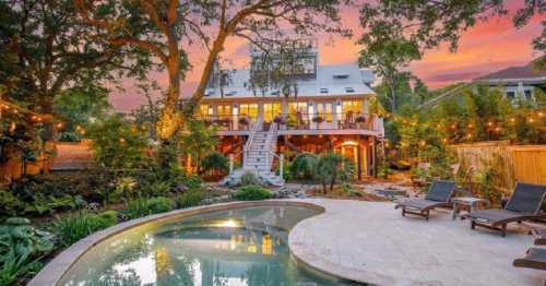 A beautiful backyard with a pool, lush greenery, and a two-story house at sunset, adorned with string lights.