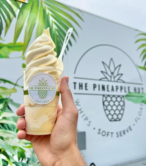 A hand holds a cup of soft serve ice cream in front of a food truck with a pineapple logo and tropical decor.