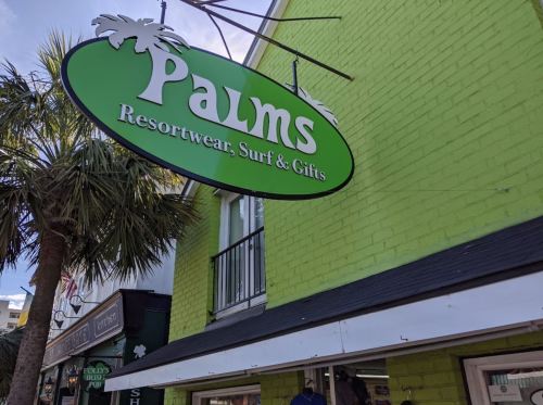 Sign for "Palms Resortwear, Surf & Gifts" on a bright green building, with palm trees nearby.