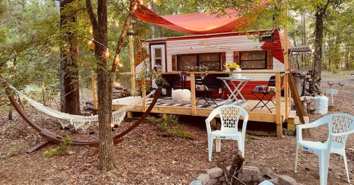 A cozy outdoor setup featuring a vintage camper, a hammock, and seating around a fire pit in a wooded area.