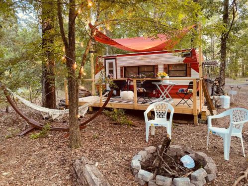Cozy outdoor setup with a vintage camper, hammock, fire pit, and seating surrounded by trees. Perfect for relaxation.