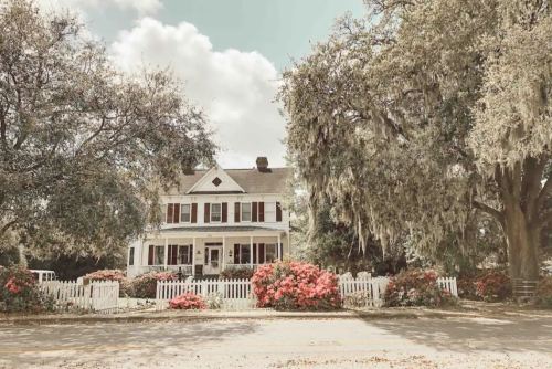 A charming white house with a porch, surrounded by lush greenery and colorful flowers, under a cloudy sky.