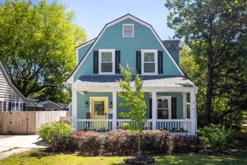 A charming blue house with a yellow door, surrounded by greenery and colorful shrubs on a sunny day.